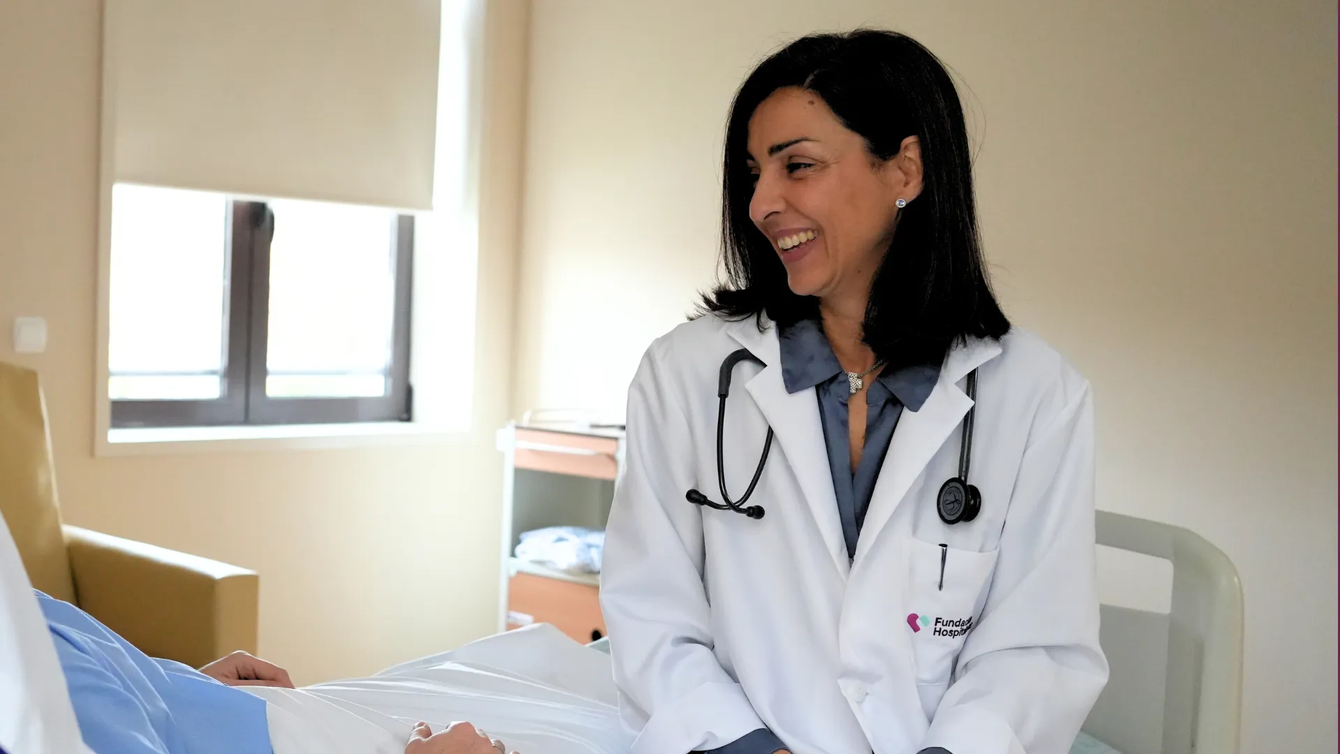 Doctora con bata blanca y estetoscopio sonríe mientras conversa con una persona ingresada en una habitación luminosa de hospital. En la bata se lee el logotipo de Fundación Hospitalarias.