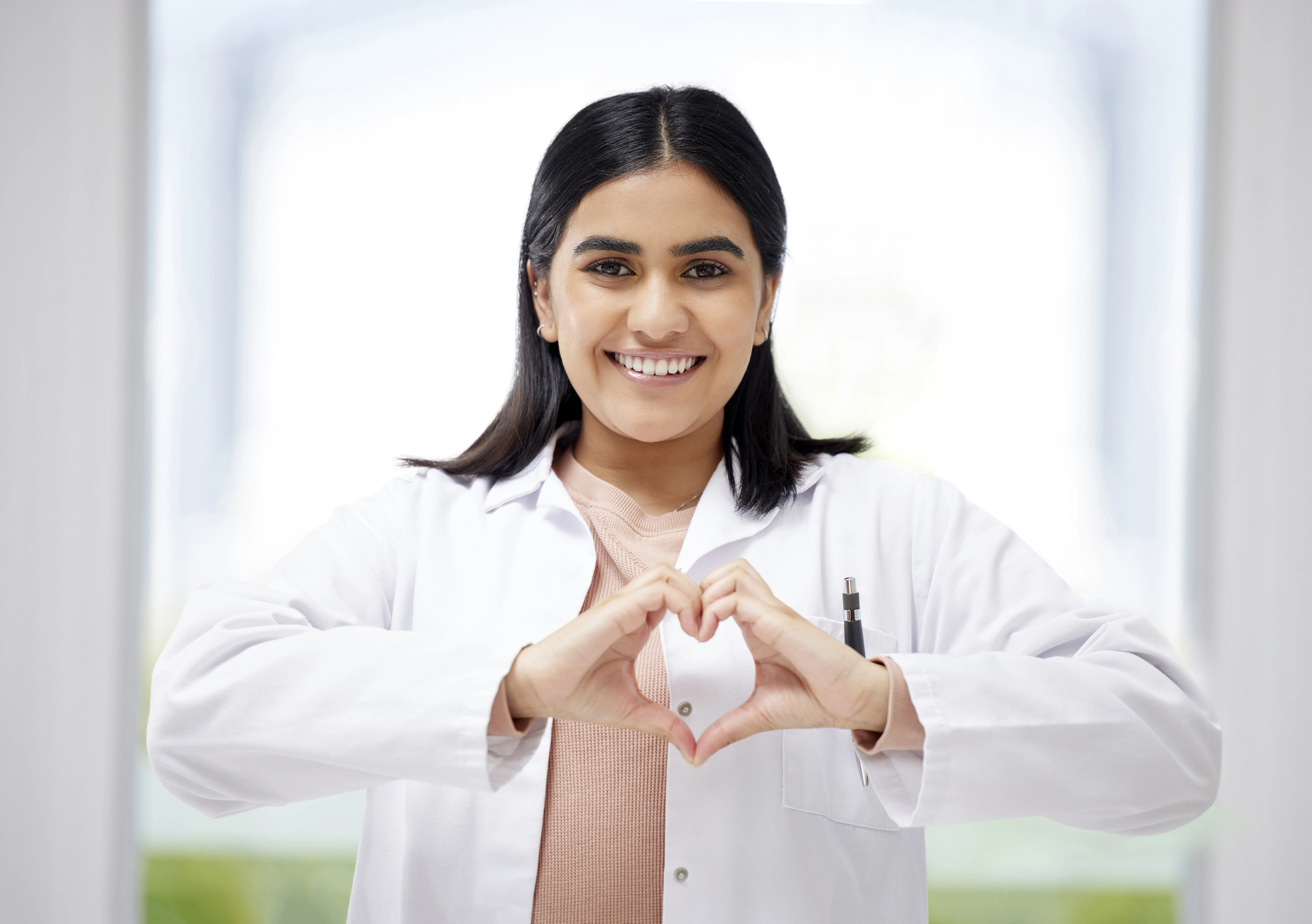 una mujer con bata blanca médica sonríe a cámara y forma un corazón con las manos