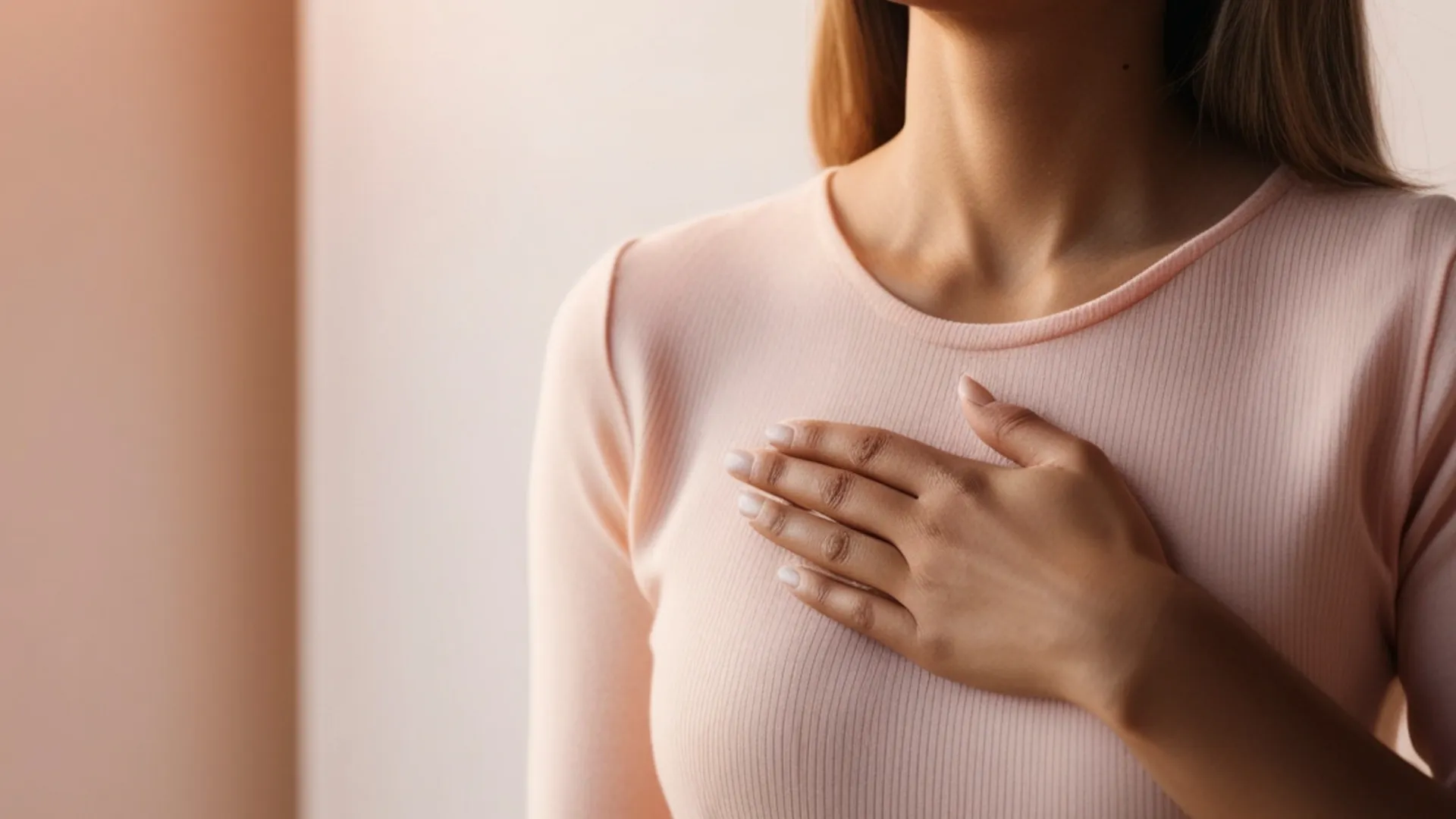 Mujer con camiseta de manga larga color rosa coloca una mano sobre la parte superior del pecho, en un gesto que indica la zona mamaria