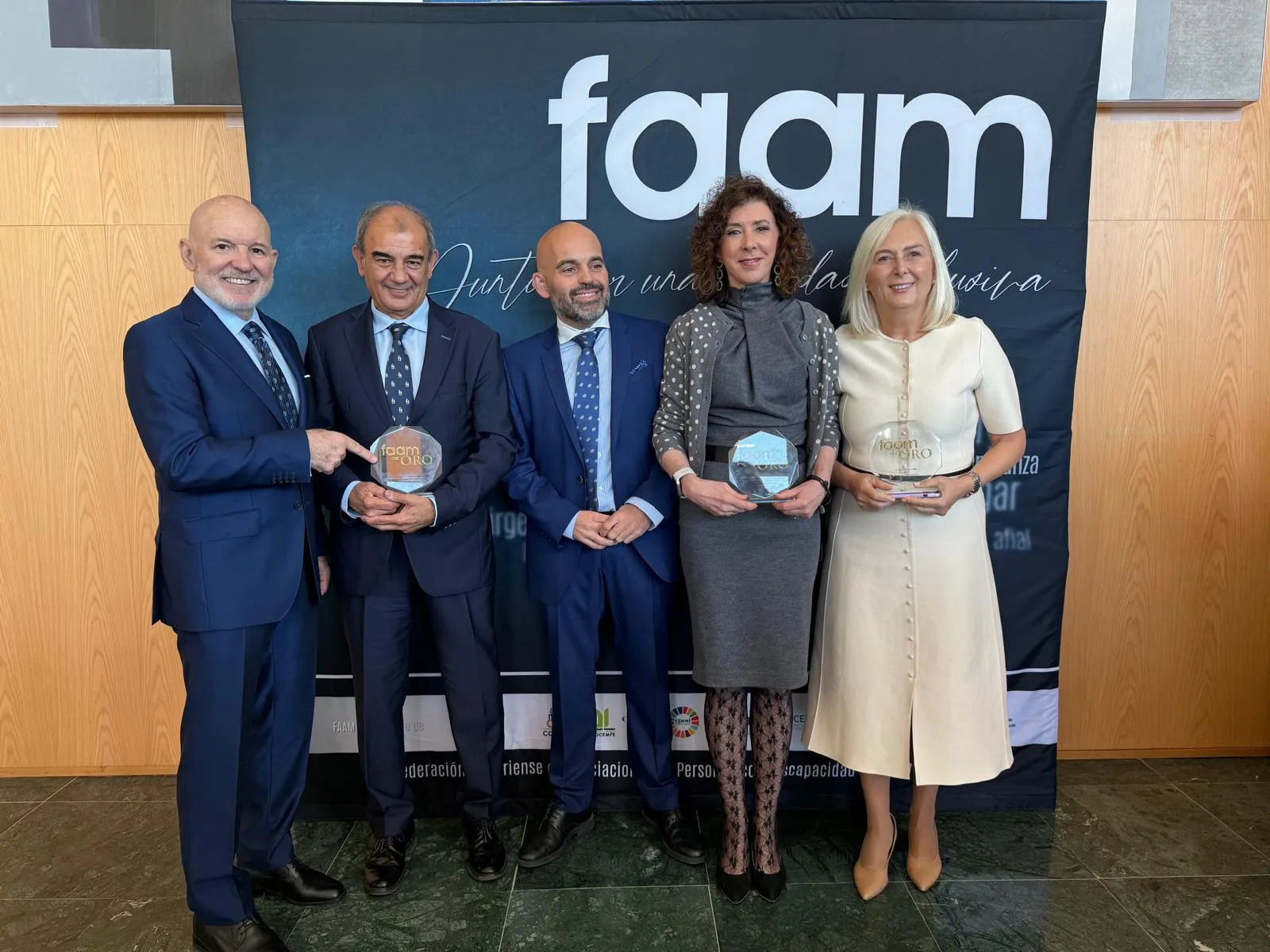 Cinco personas vestidas de manera formal posan frente a un panel con el logotipo de FAAM y el lema 'Juntos una vida activa'. Varias de ellas sostienen trofeos circulares transparentes, indicando que se trata de una ceremonia de entrega de premios o reconocimiento.