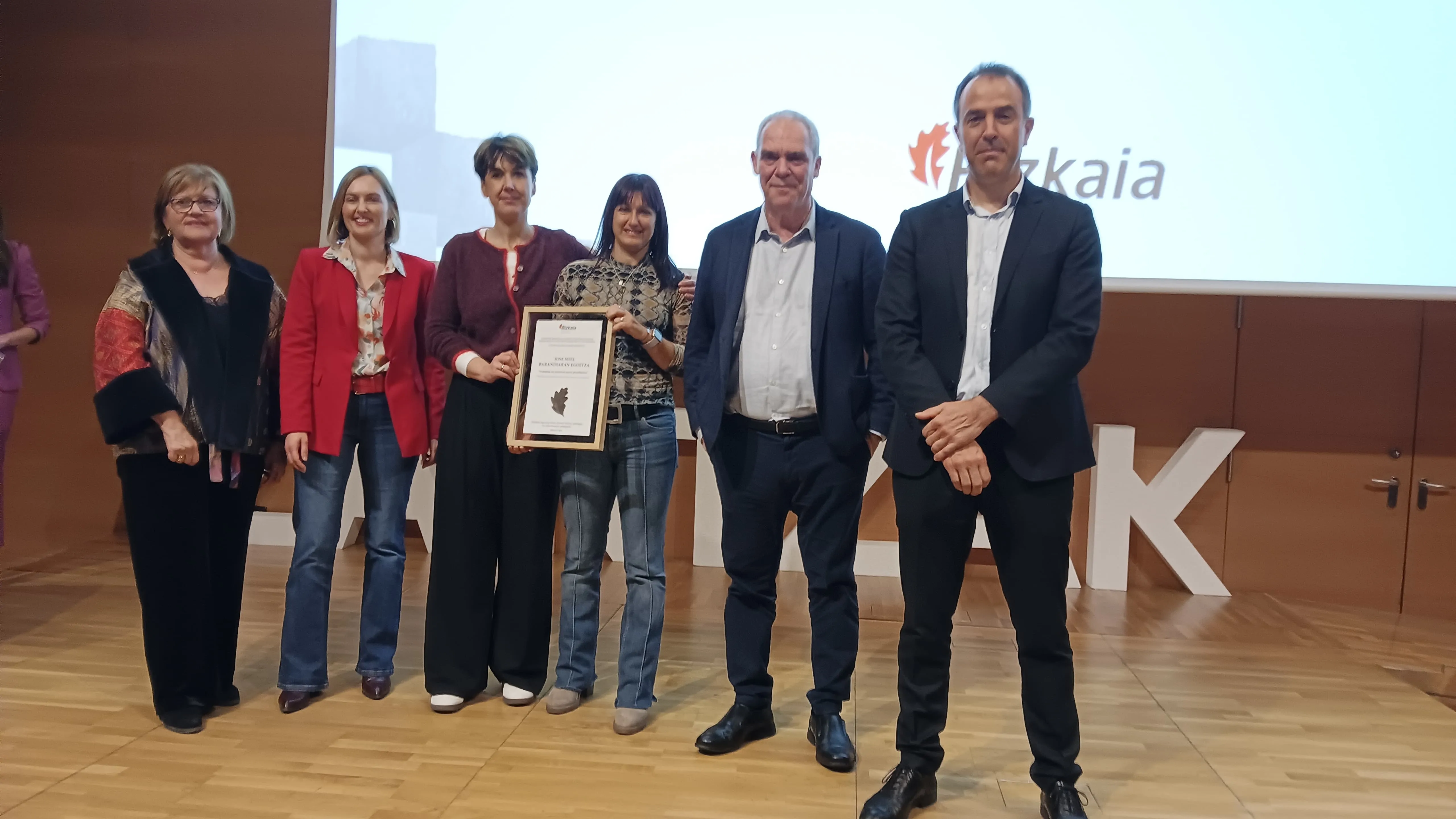 Grupo de cinco personas posando en un escenario de madera, sosteniendo un diploma enmarcado. Al fondo se observa una pantalla blanca con el logotipo de Bizkaia