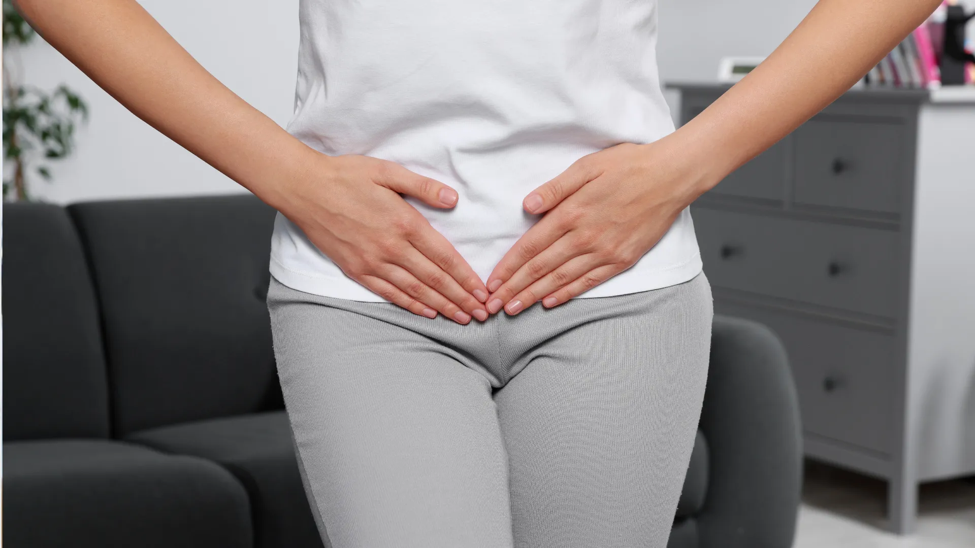 Persona vestida con camiseta blanca y pantalón gris coloca ambas manos sobre la zona inferior del abdomen