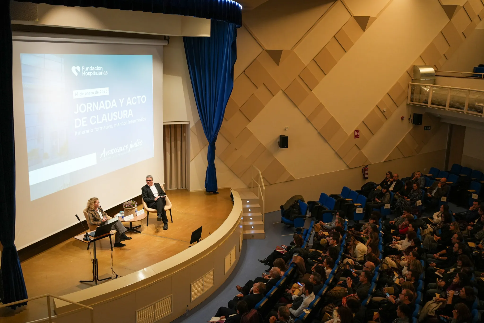 dos personas en un escenario. a la derecha se ve el público asistente durante la jornada de clausura del itinerario de liderazgo para mandos intermedios de la Fundación Hospitalarias