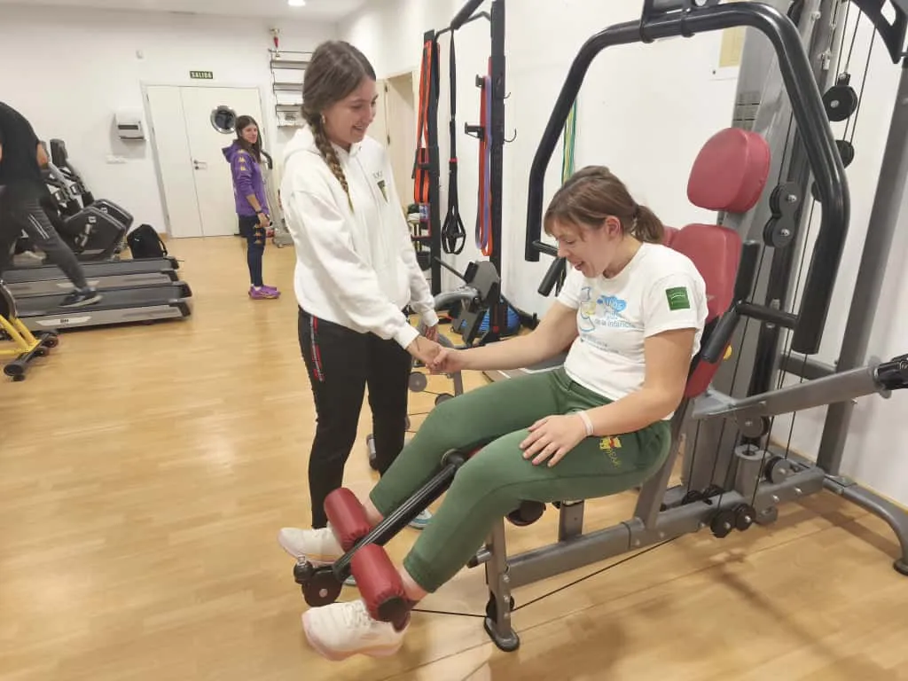 voluntaria en una sala de rehabilitación donde una persona usuaria está ejercitándose en una máquina de cuádriceps