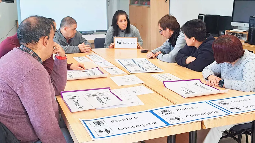 Grupo de personas participando en una actividad sobre accesibilidad cognitiva. Están sentados alrededor de una mesa con varios carteles y señales que indican diferentes plantas y ubicaciones, como 'Planta 0', 'Planta 1', 'Conserjería'. En el centro de la 
