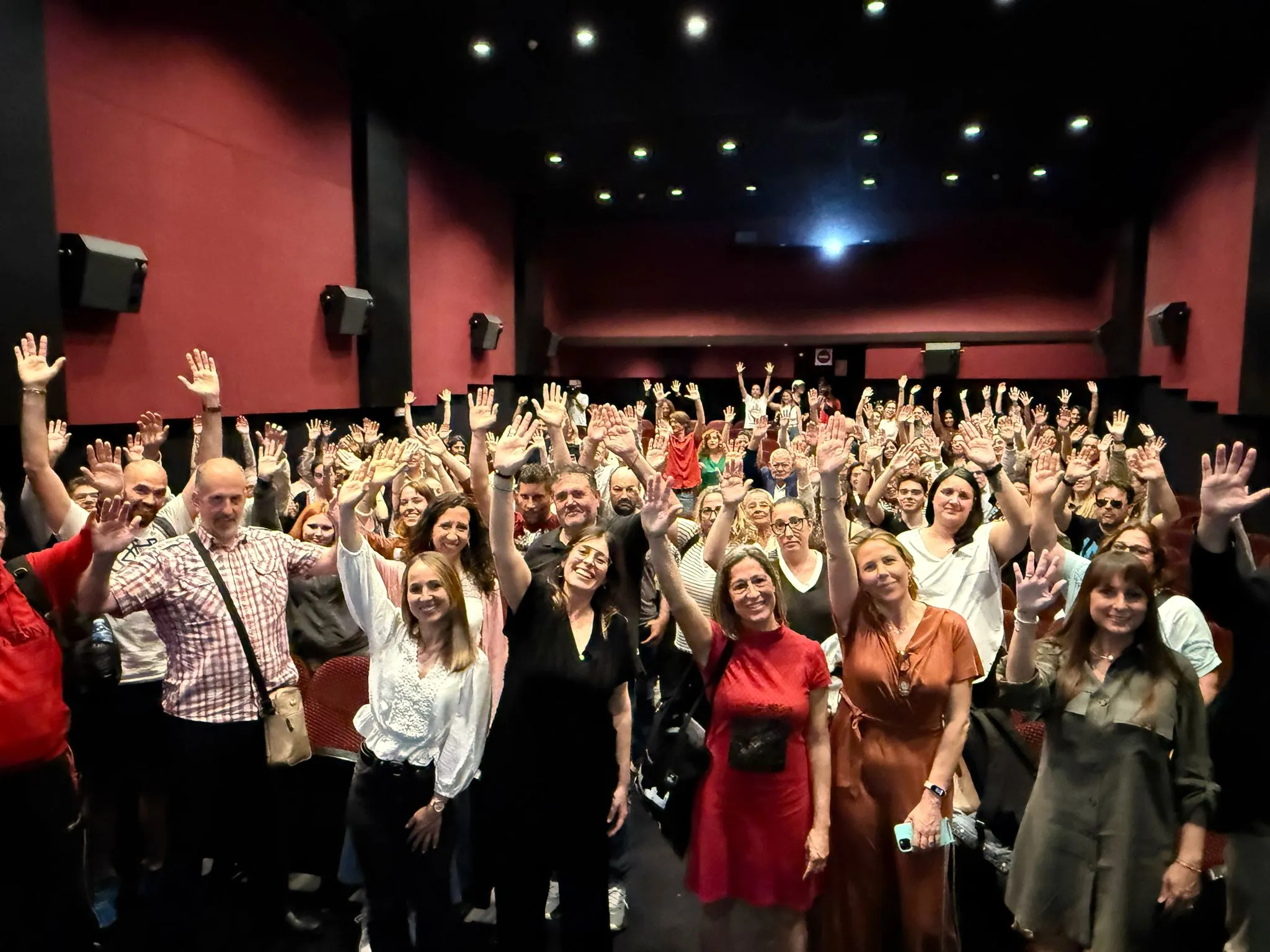 Grupo numeroso de personas en una sala de cine o auditorio, de pie y con las manos levantadas, sonriendo hacia la cámara, en un ambiente festivo y participativo. preestreno votemos en barcelona fundacion hospitalarias