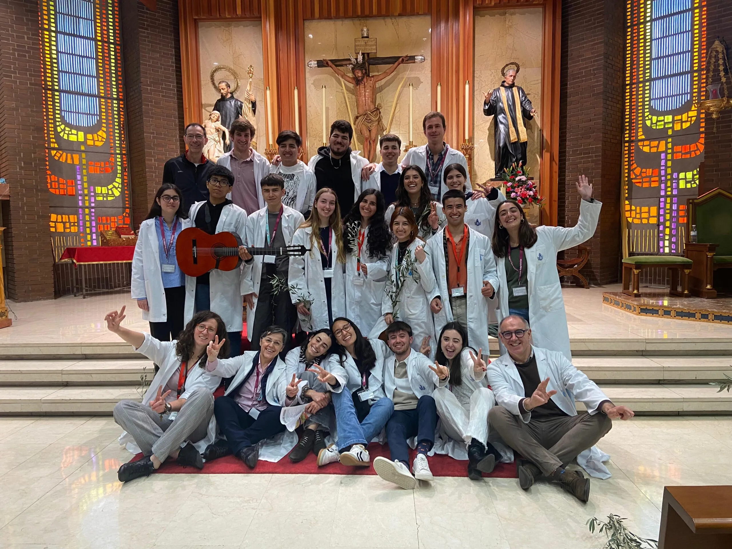 Grupo de jóvenes con batas blancas posando sonrientes en el interior de una iglesia, junto a personal adulto. Al fondo, un altar con figuras religiosas y vitrales de colores.