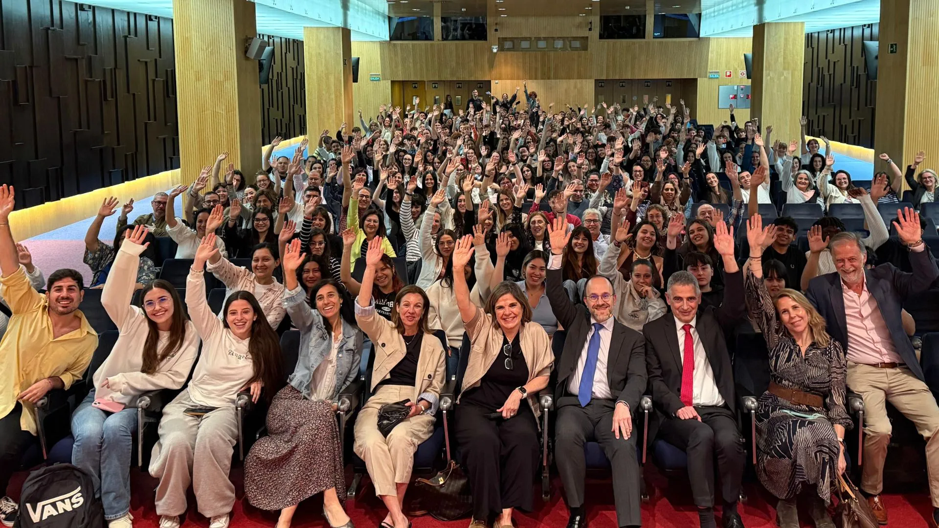 Gran grupo de personas sentadas en un auditorio durante el preestreno de la película Votemos en Zaragoza, levantando la mano y sonriendo hacia la cámara. En primera fila, varias personas con traje y corbata. El ambiente es alegre y participativo. Organiza