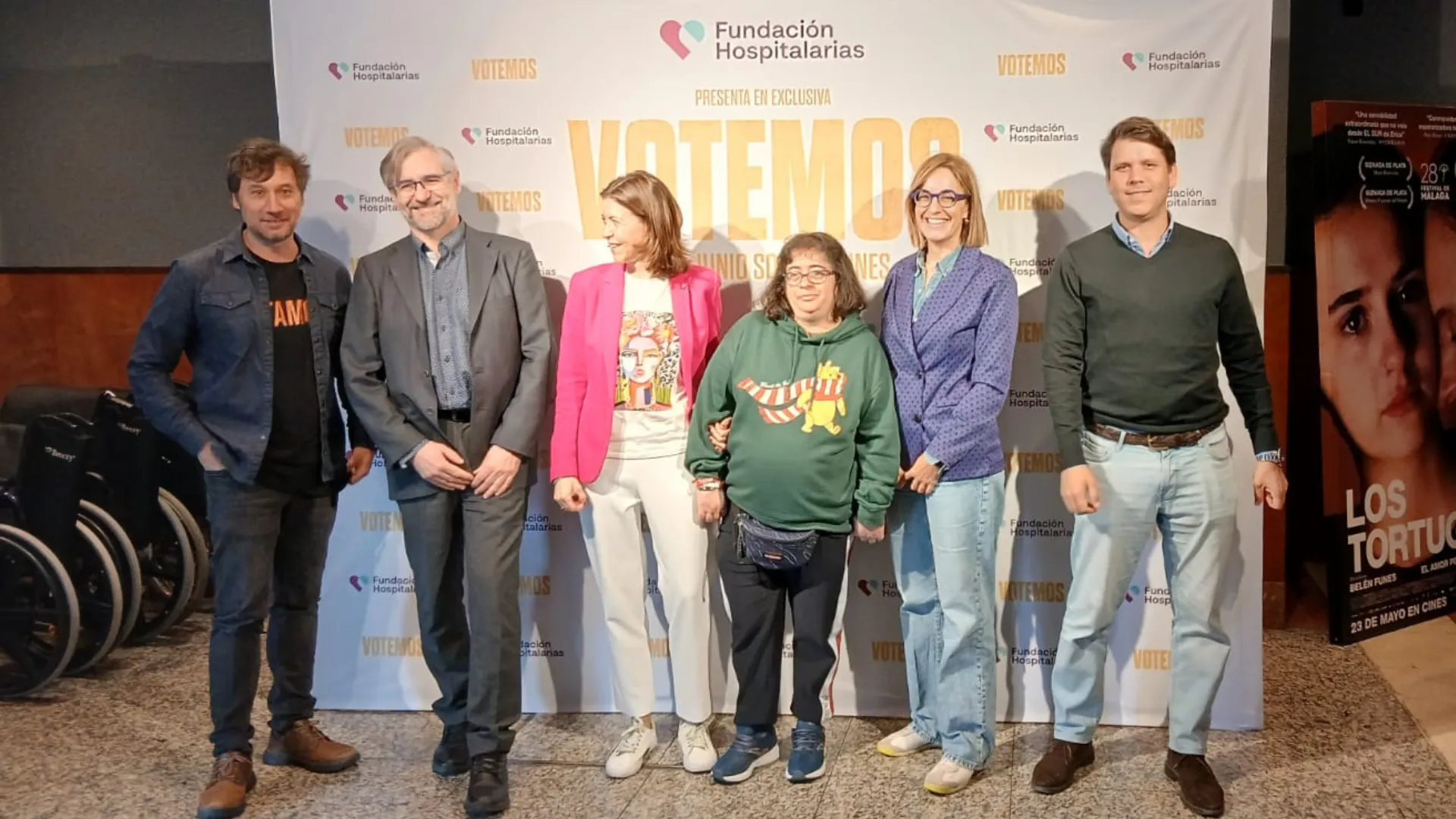 Grupo de seis personas posando frente al photocall del preestreno de 'Votemos' en Fundación Hospitalarias Pamplona. El fondo muestra el logo de la Fundación y el título del cortometraje. Al fondo, se ven varias sillas de ruedas.